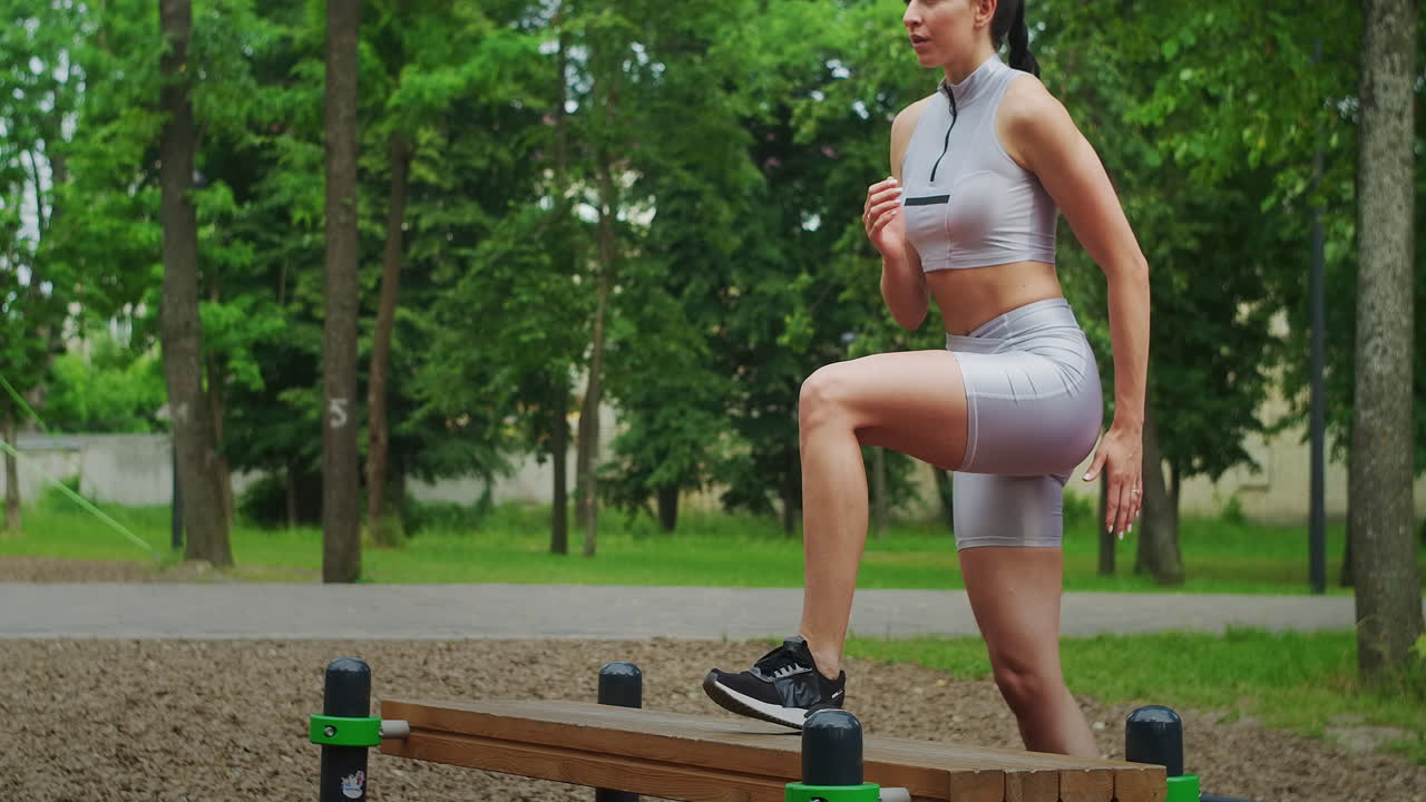 una mujer joven en un parque realiza ejercicios caminando en un banco con ropa deportiva en el verano.
