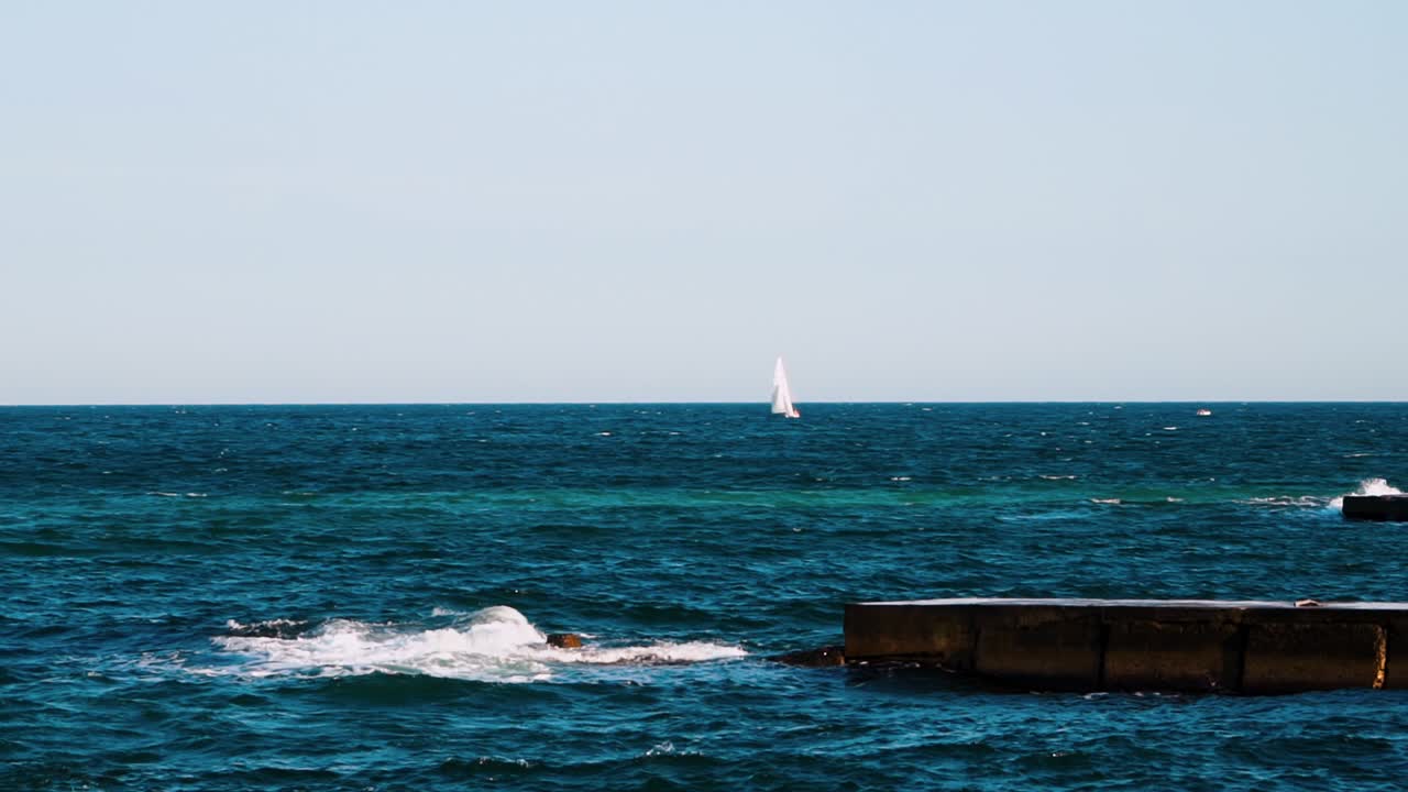 Blue water background. Stone dock in the sea water. White sail floating in the sea far away. Waves breaking on pier and making white foam. Slow motion.