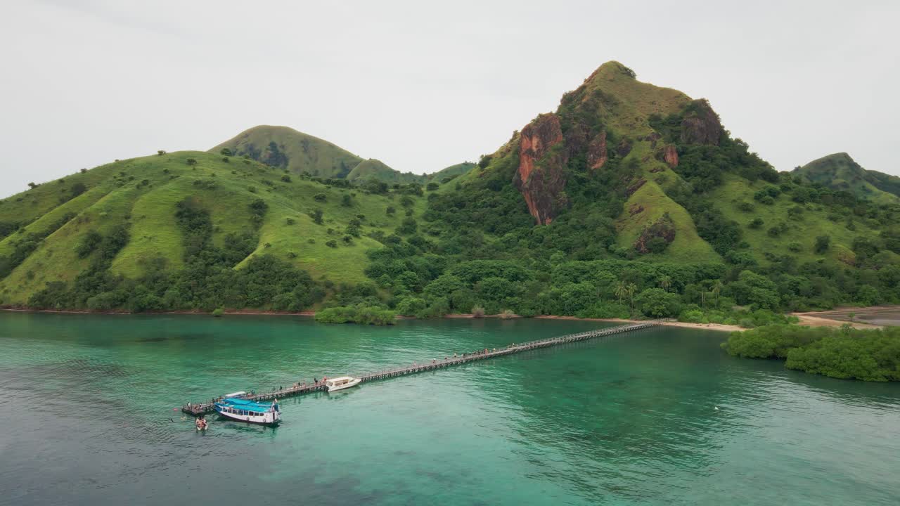 The dramatic view of rugged hills stand above the green emerald waters of Komodo National Park, Indonesia with a wooden pier reaching out into the crystal-clear ocean.
