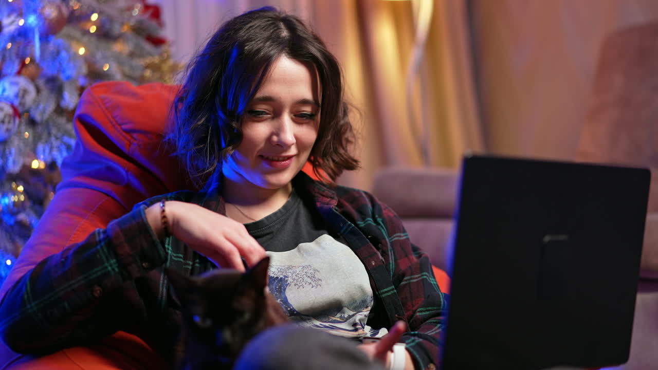 Relaxed Caucasian female in checkered shirt sits using a laptop in a chair. Woman works remote from home petting a cat.