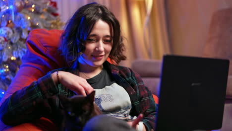 Relaxed Caucasian female in checkered shirt sits using a laptop in a chair. Woman works remote from home petting a cat.