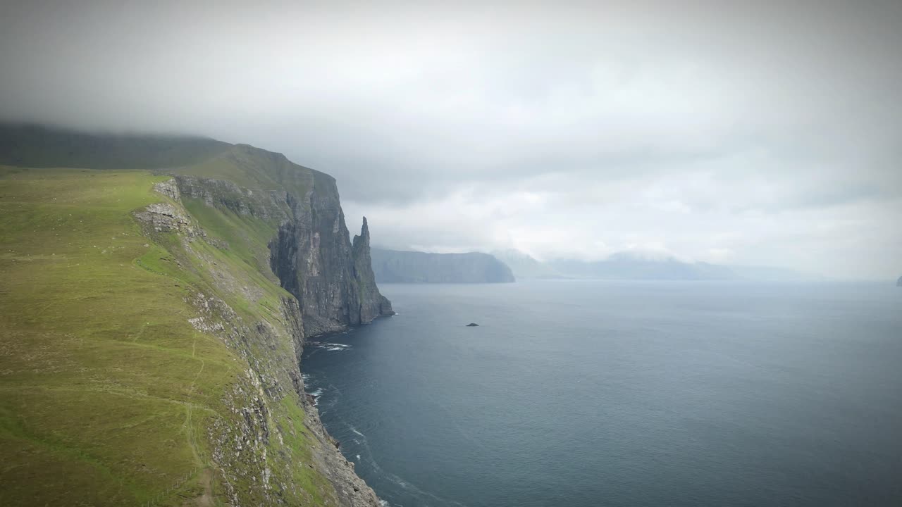 tomada aérea de trøllkonufingur, una pila de mar que se asemeja al dedo de una bruja, que se eleva desde el océano cerca de la costa de sandavágur, vágar, islas feroe, bajo un cielo nublado