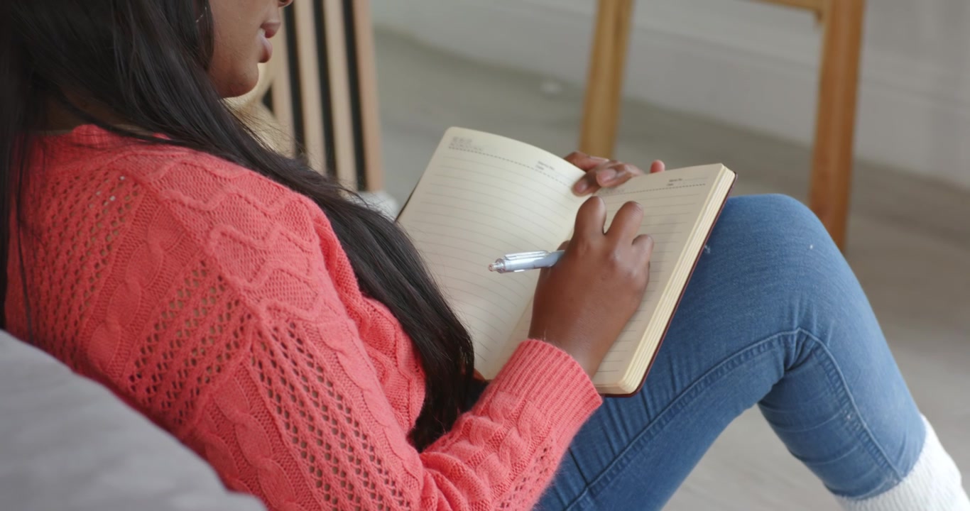 Donna afroamericana che si sistema sul pavimento accanto al divano scrivendo in un'agenda con una penna, cuffie e tazza accanto