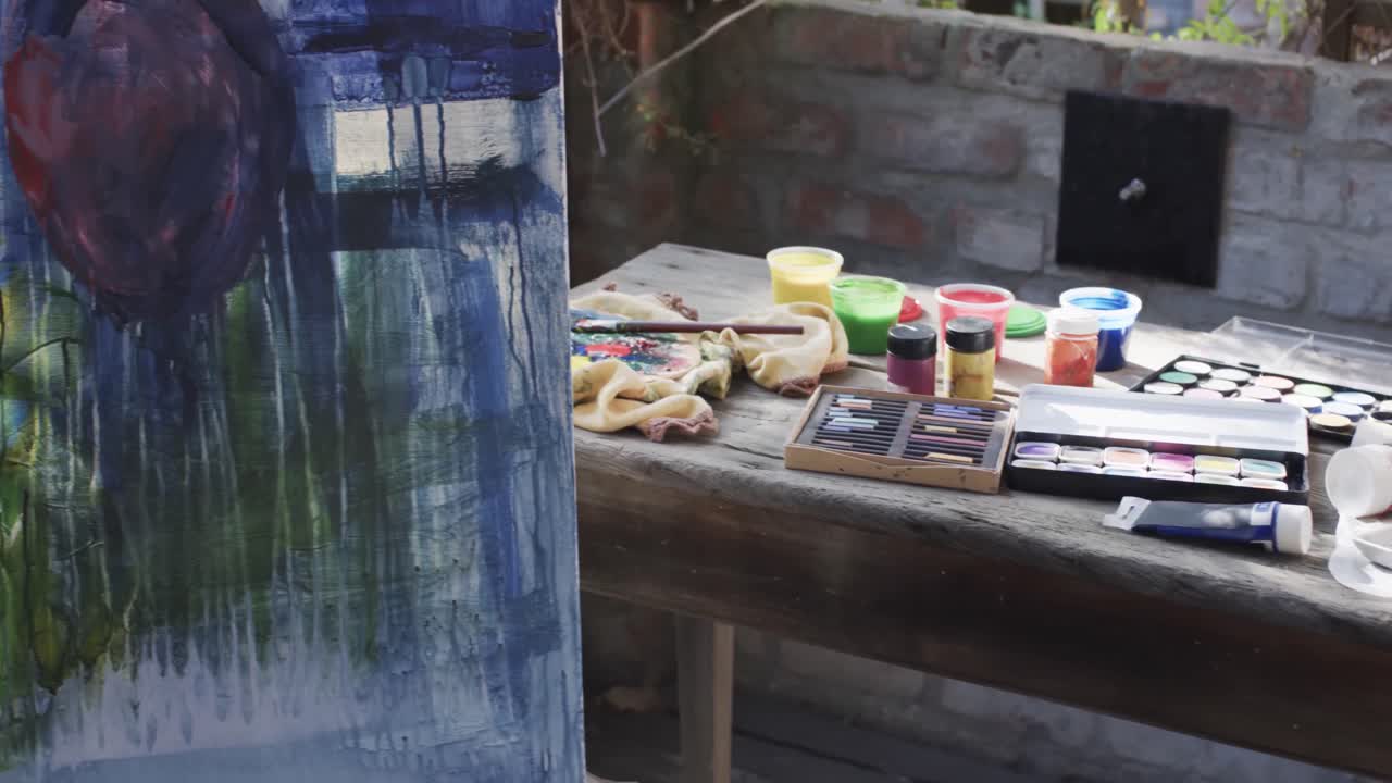 Close up of painting on easel and paints and brushes on table in sunny garden, slow motion