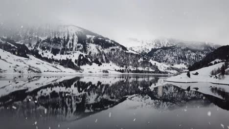 Montañas-De-Invierno-Reflejo-Del-Agua-Del-Lago-Durante-Las-Fuertes-Nevadas,-Vista-Aérea-De-Drones