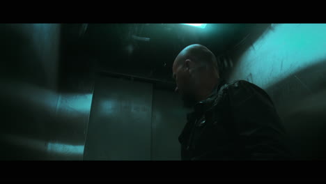 a bald dangerous looking man prepares a gun with the silencer in the elevator