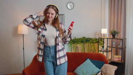 overjoyed young woman in wireless headphones dancing, singing on cozy couch in living room at home