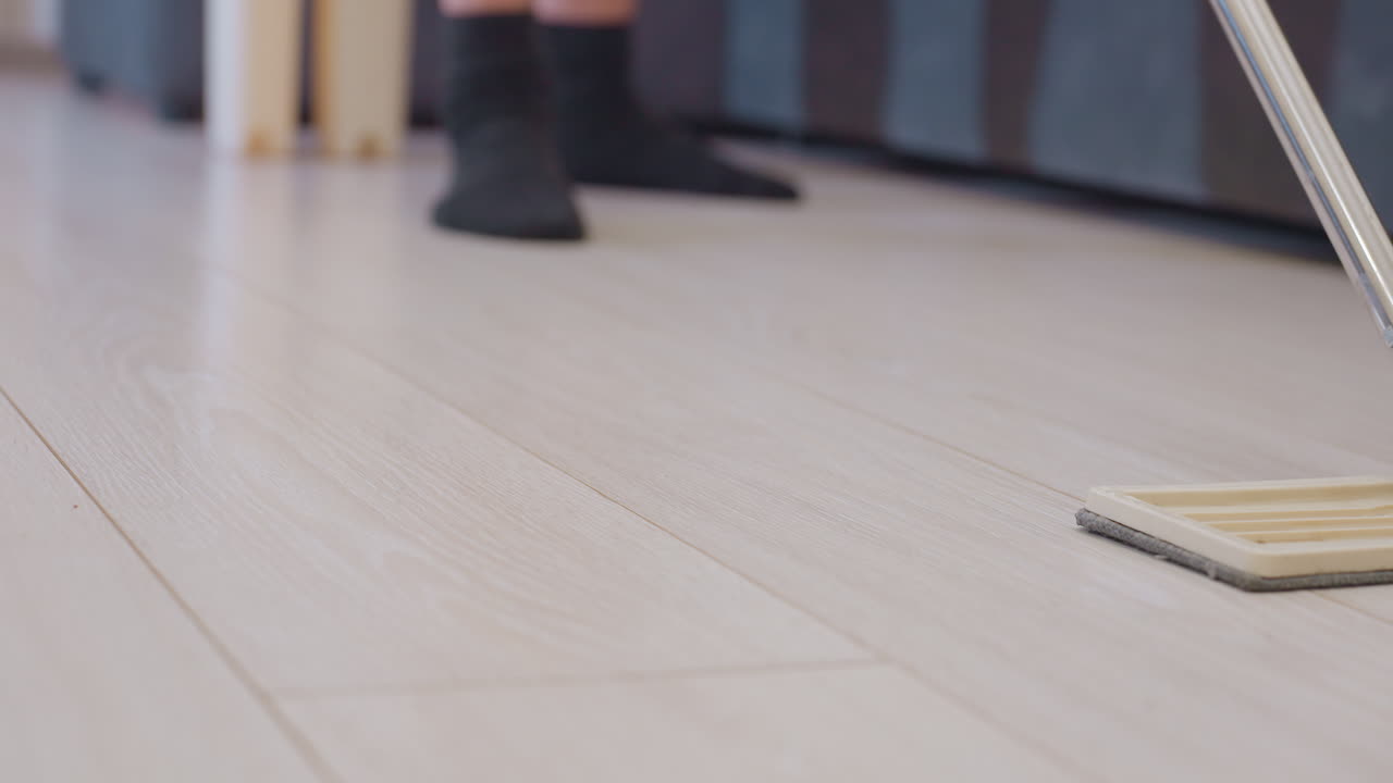 Lower angle of adult pushing mop across wooden floor near sofa while wearing black socks showing detailed household cleaning task and daily domestic routine