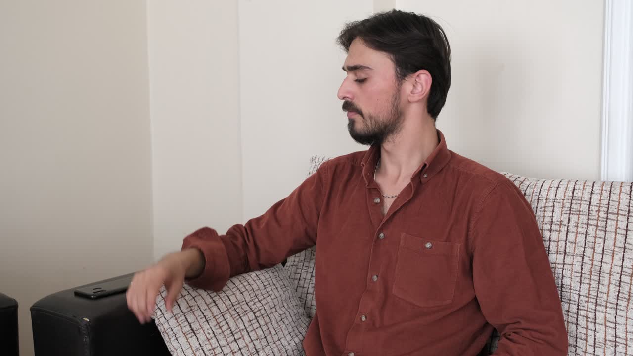 The Caucasian man sitting in the house on a sofa, or couch and using a mobile phone, the young man playing with his beard and waiting to pick up the phone