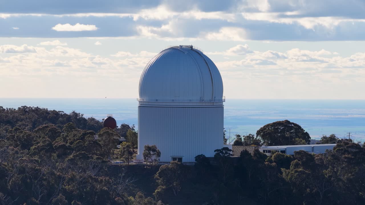 Drone camera slowly pans across a large white observatory dome surrounded by dense forest, under soft daylight with partly cloudy skies and distant horizon