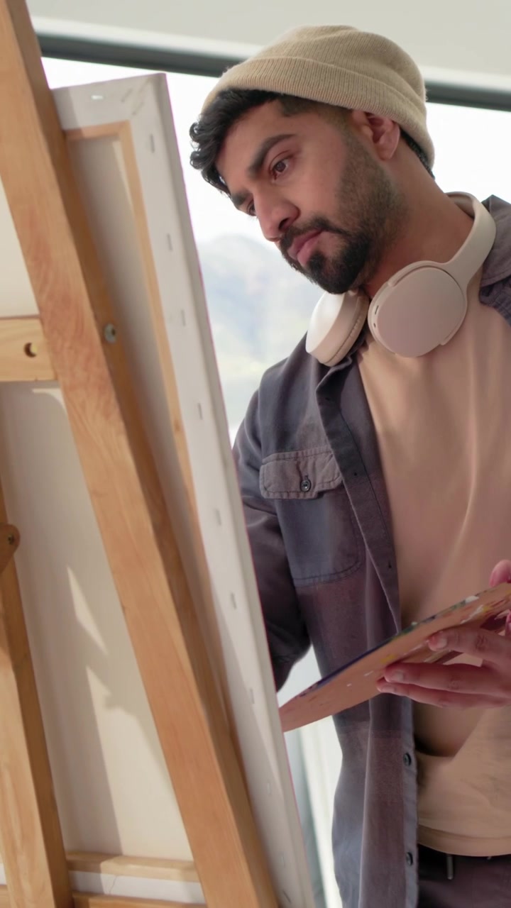 Video vertical: Artista contemplando el lienzo, mezclando pintura y pintando, utilizando la vista de la ventana, un pincel y un gorro.