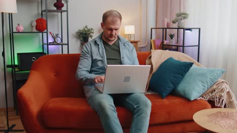 caucasian man sitting on sofa opening laptop pc starting work elearning online in room at home