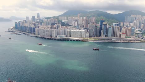 Convoy-of-local-Fishing-boats-causing-in-Hong-Kong-Victoria-bay,-with-city-skyline-in-the-horizon,-Aerial-view