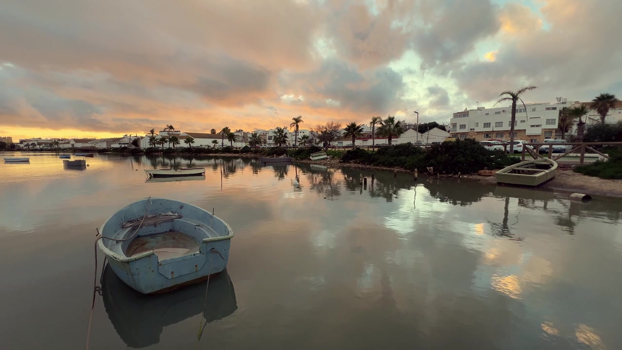 Små fiskerbåde flyder på roligt vand i Barbate, Andalusien, Spanien, under en fantastisk solnedgang, med farverig himmel der spejler sig i vandet og hvide bygninger i nærheden.