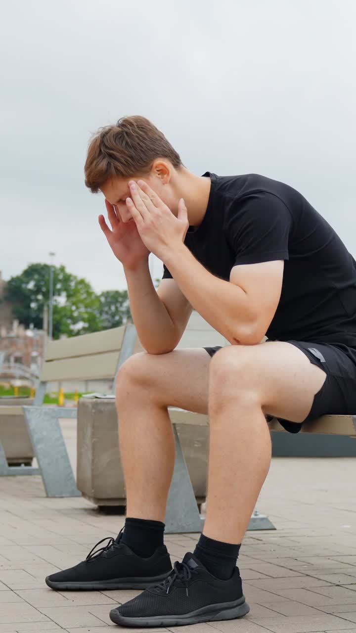 Man in black shirt sits on a bench, covering face with hands, feeling down, vertical