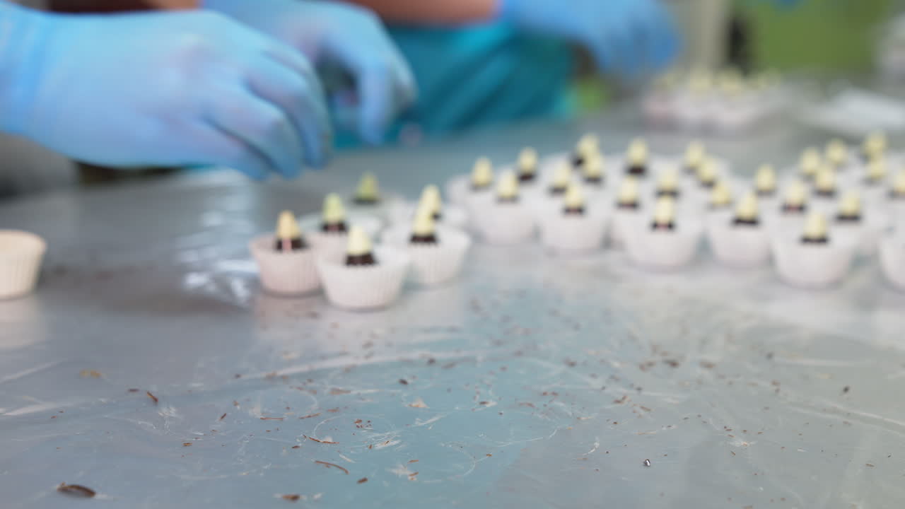 Confectioner takes little chocolate cones with white tops and puts them in paper. Packaging ready-made sweets at factory. Blurred backdrop.