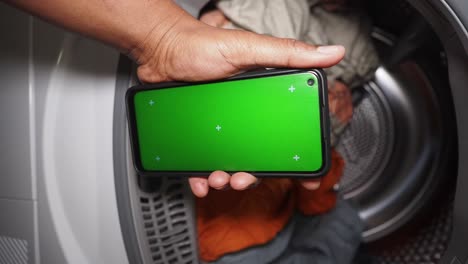 Hand holding a smartphone with green screen in front of a laundry machine