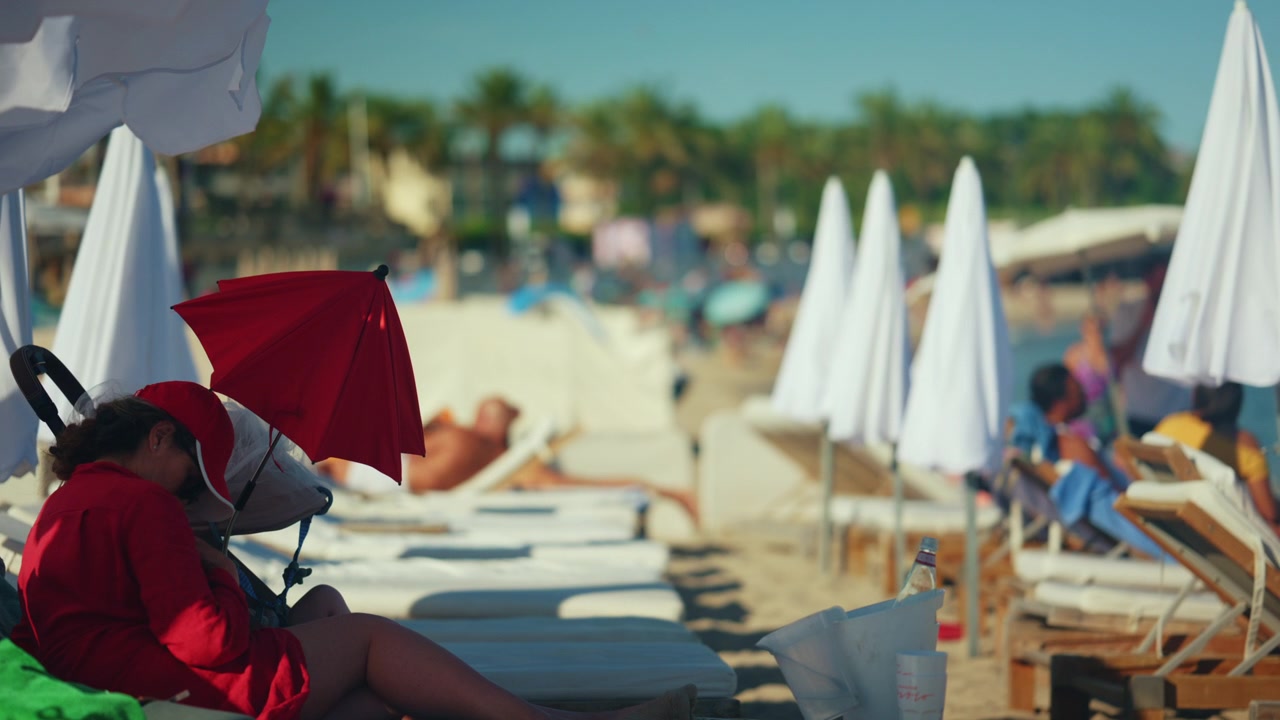 Momento familiare in spiaggia con un bambino seduto vicino a un passeggino che tiene un ombrello rosso acceso, in contrasto con i toni neutri e delicati della spiaggia, Antibes, Francia.