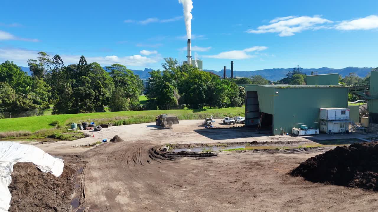 Tractor and bulldozer work near a factory with smoke stacks under clear skies, highlighting industrial activity in a rural landscape