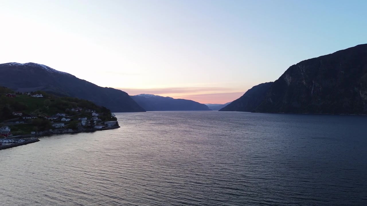 Skumringens blide skær oplyser en rolig fjord i Norge, indrammet af fjerne sneklædte tinder. Velegnet til rejse- eller landskabsprojekter.