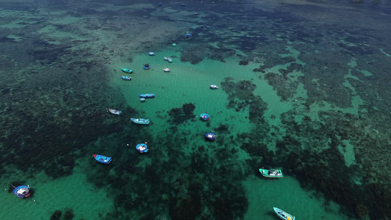 Dynamic drone forward shot capturing vibrant fishing boats anchored above a coral reef in Ninh Hải District during bright midday light.