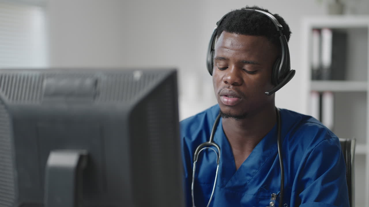Medico o infermiere afroamericano con cuffie e computer che lavora in ospedale. Giovane medico o terapista professionista che consulta un paziente utilizzando la comunicazione a distanza e parlando in webcam