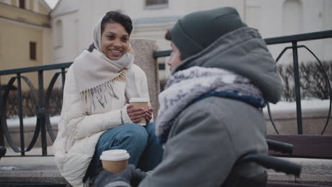 Mujer-Musulmana-Y-Su-Amiga-Discapacitada-En-Silla-De-Ruedas-Bebiendo-Café-Para-Llevar-En-Un-Banco-En-La-Ciudad-En-Invierno-3