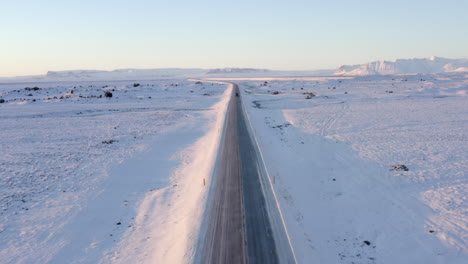 AERIAL:-Snow-White-Landscape-with-Road-following-Jeep-in-Iceland-at-Sunset-Winter,-Sun,-Arctic