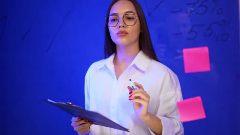 Calm confident lady in glasses writes with marker on the glass board. Portrait of a business lady making financial presentation.