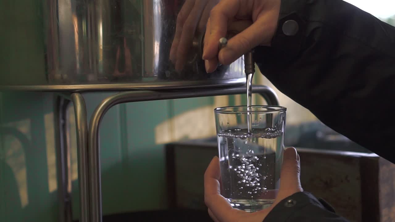 A lady is pouring water from a metal water tank to a tall glass in slow motion.