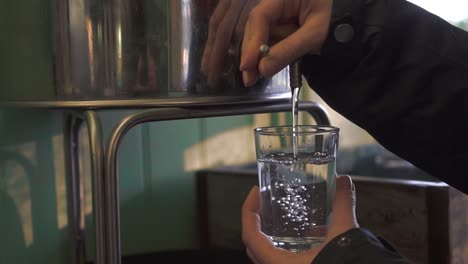 a lady is pouring water from a metal water tank to a tall glass in slow motion.