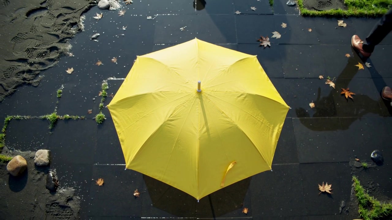 Ombrello giallo che si tende con uno scatto su piastrelle bagnate, due adulti con pantaloni scuri camminano sulle pozzanghere.