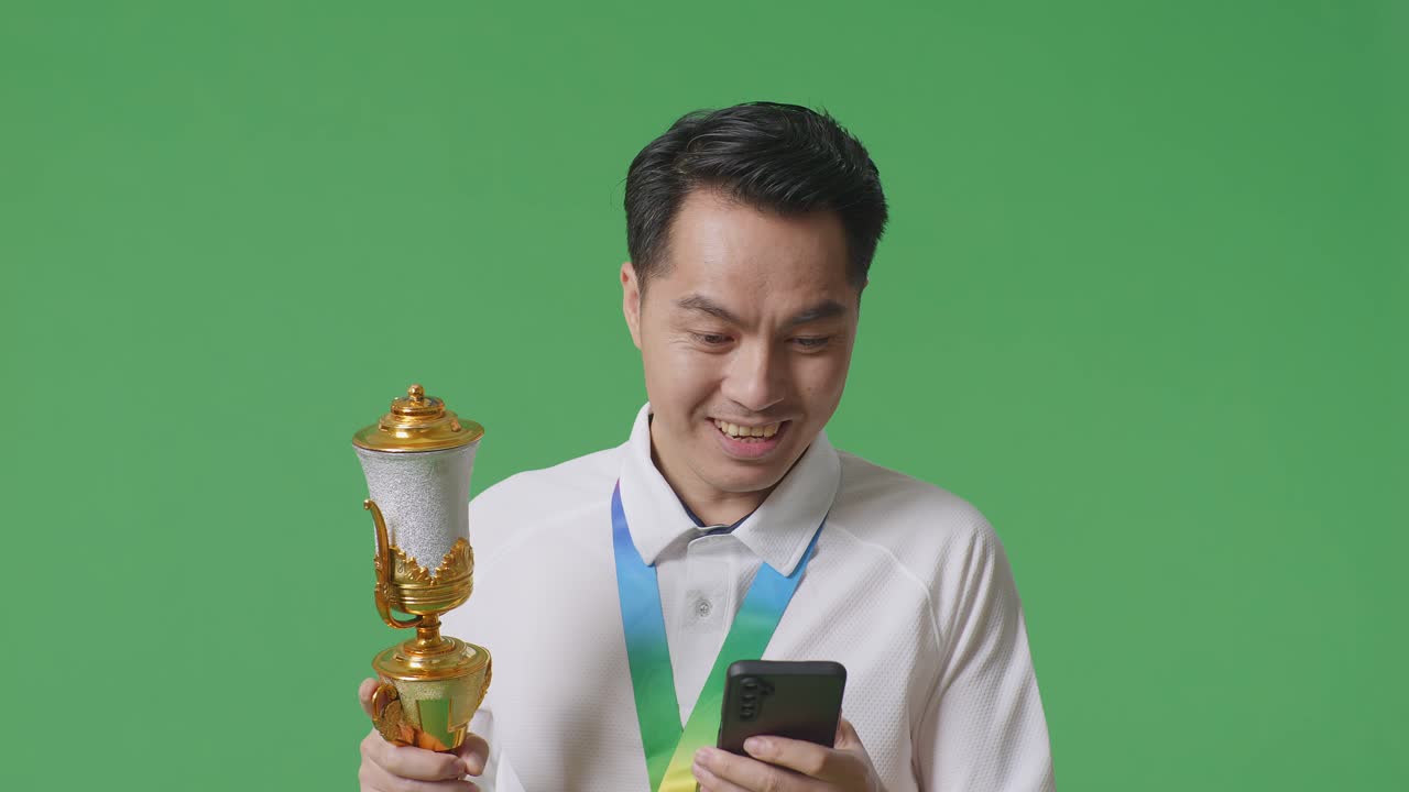금메달과 트로피를 들고 있는 아시아인 남자가 스마트폰을 쳐다보며 스튜디오의 녹색 스크린 배경에서 와우라고 말합니다.