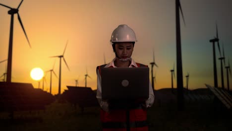 asian female engineer in a helmet standing in front of wind turbines rotating at sunset, working on a laptop and looking around