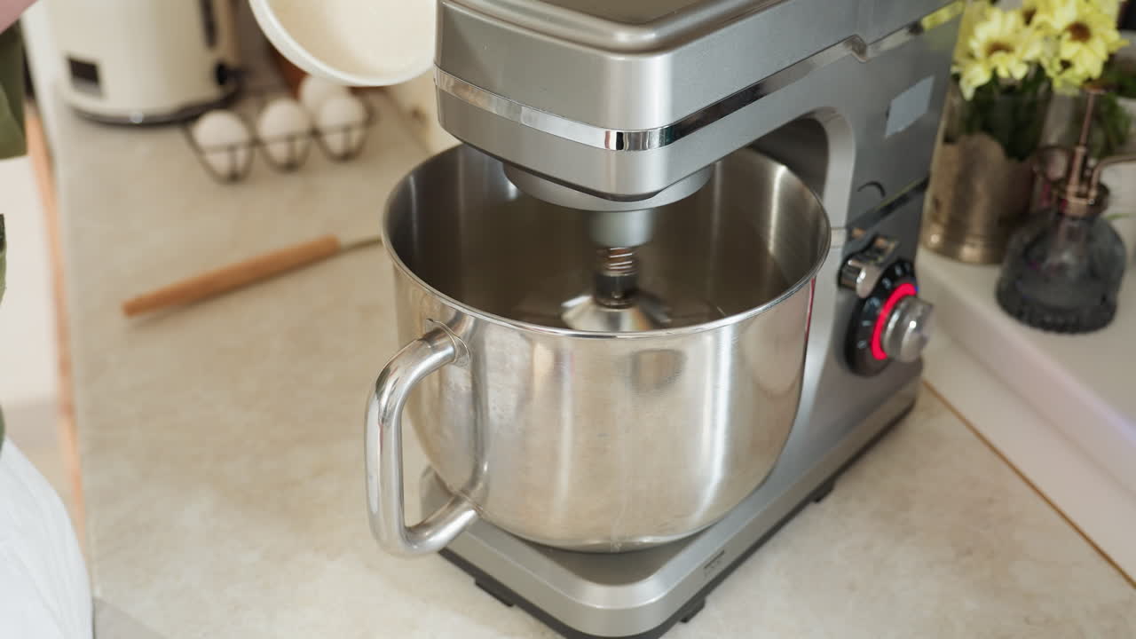 Close up of stainless steel electric mixer in action on kitchen counter while person prepares to pour flour from white bowl into rotating bowl during home baking in clean