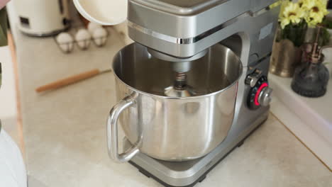close up of stainless steel electric mixer in action on kitchen counter while person prepares to pour flour from white bowl into rotating bowl during home baking in clean