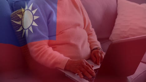 animation of flag of taiwan waving over midsection of asian senior woman using laptop on sofa