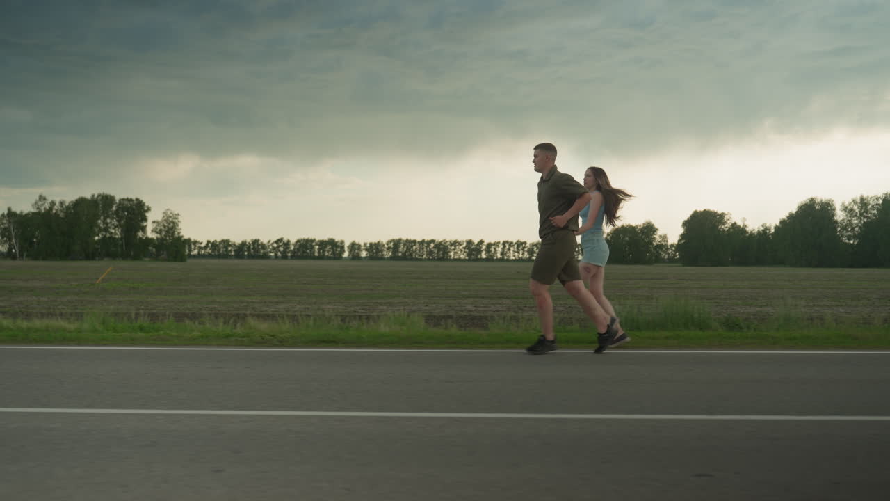 side view of man and woman running hand in hand along rural road edge under dramatic cloudy sky at golden hour, her long hair flowing, his khaki outfit contrasting green fields and asphalt road