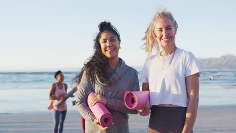 group of diverse female friends holding yoga mats at the beach