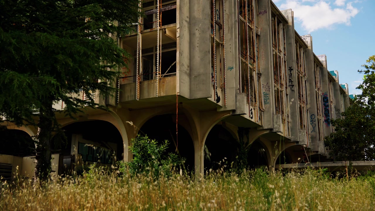 Edificio di un hotel abbandonato circondato da erba alta e alberi. Struttura in cemento vuota con graffiti, finestre rotte e segni di abbandono in un ex resort turistico.