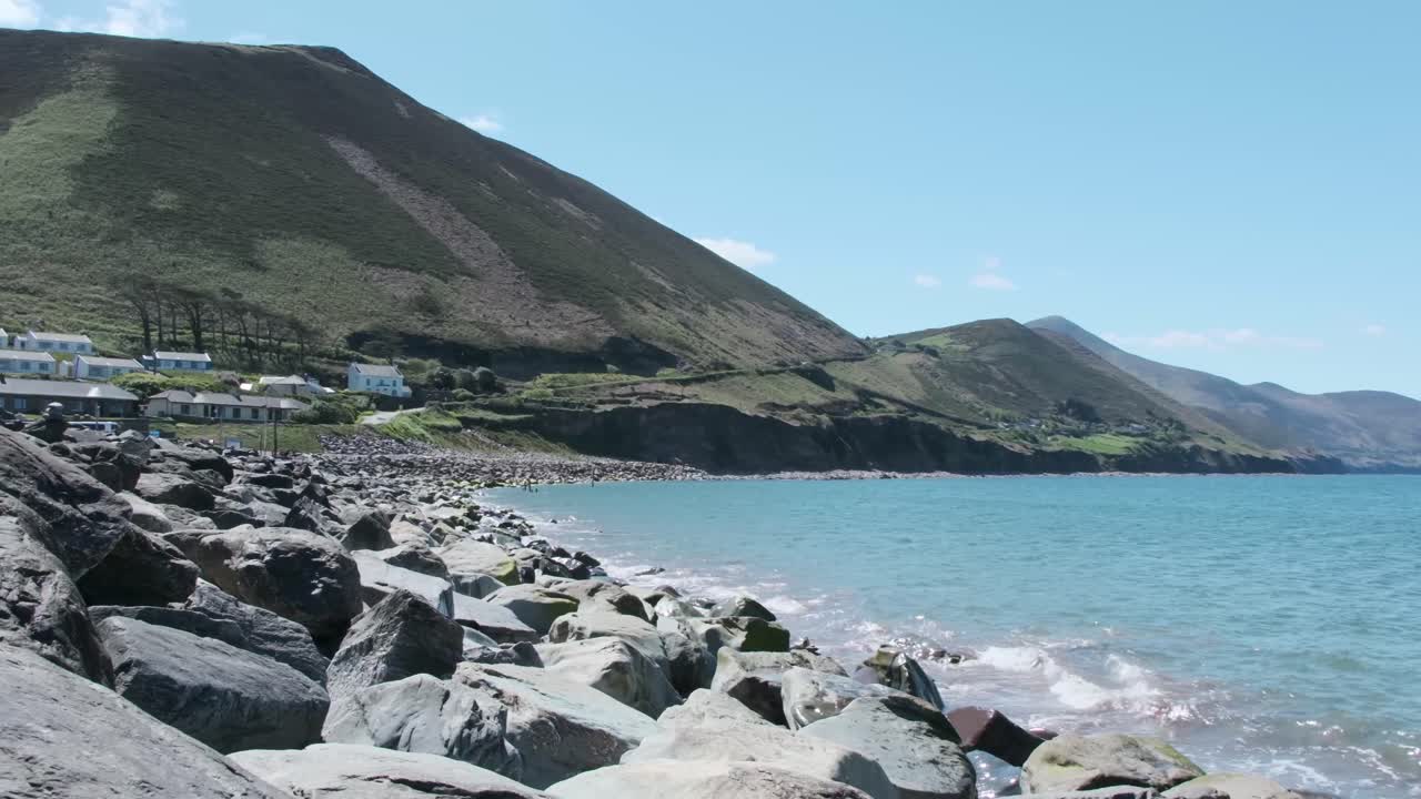 Handheld shot from rocky shore in Rosbeigh, Ireland. Gentle waves meet the coast with hills rising behind. Calm, natural beauty on a peaceful summer day.