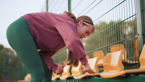 side view of athlete cleaning yellow stadium seat with wiper, holding something in left hand while her hair cascades down, outdoor field with stadium seating and green fence in background