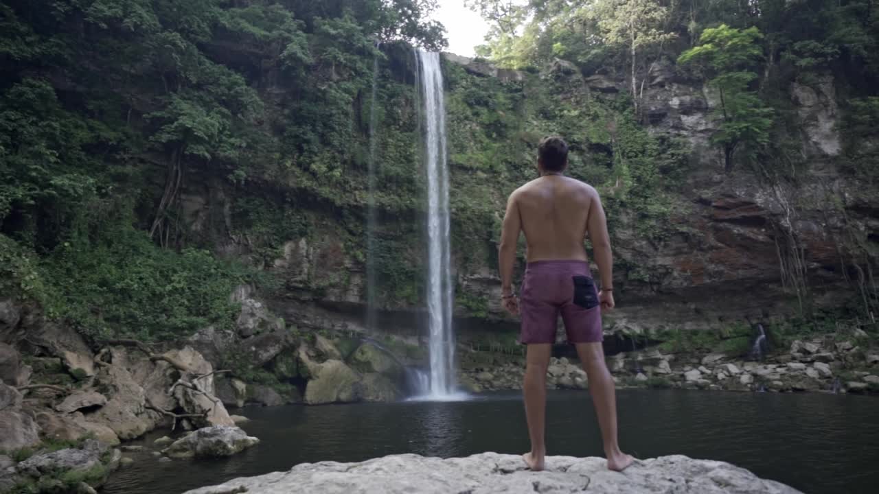 A man, viewed from behind, stands on a rock platform at the base of the Misol-Há waterfall At sunrise in Chiapas, México