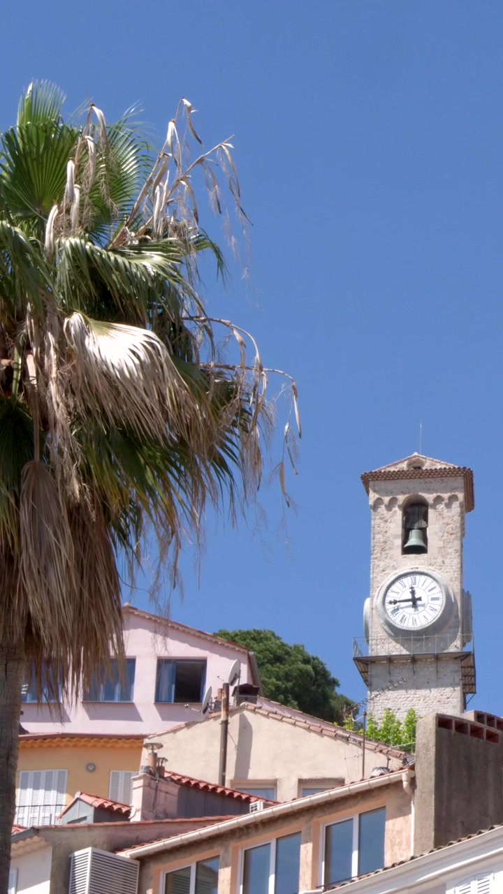 Inquadratura statica di una torre dell'orologio in pietra sopra edifici tradizionali sotto un cielo azzurro e limpido.