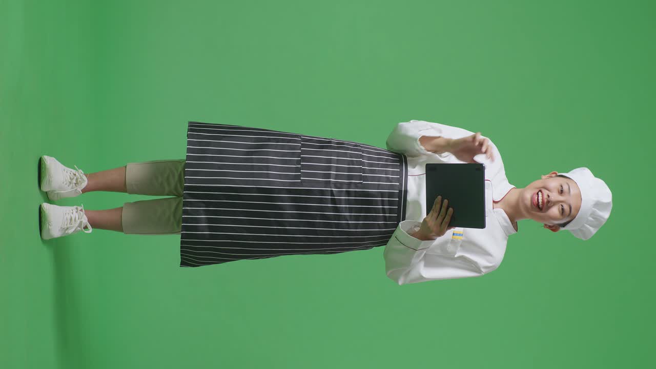 Full Body Of Asian Woman Chef Using A Tablet And Pointing To Side While Standing In The Green Screen Background Studio