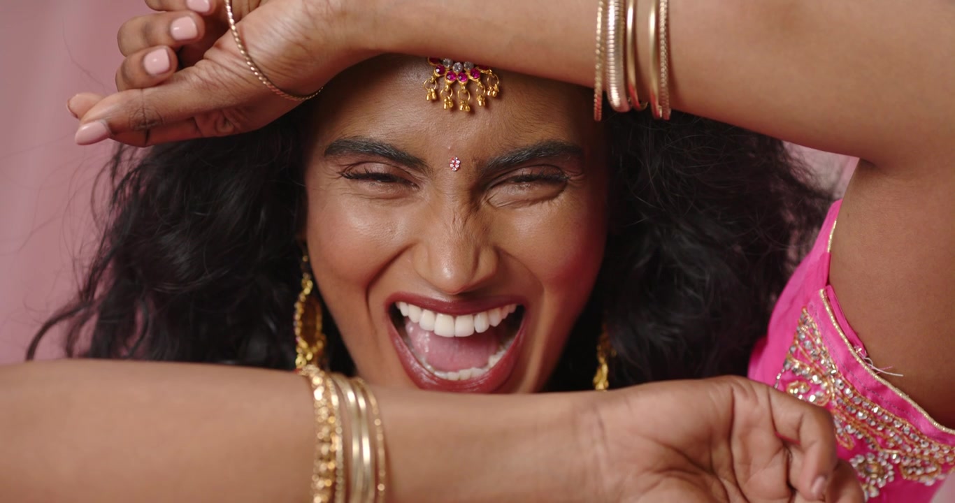 Indian woman reacting to camera, smiling, lifting arm, peeking through gold bangles at studio