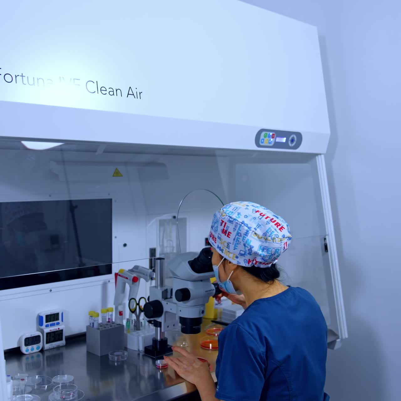 Rear view of the lab technician sitting and looking in microscope. Medic looks carefully at Petri dish in her hands