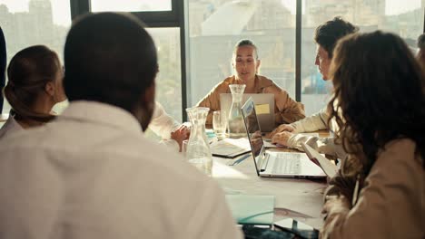 Offline-conference-of-office-workers,-a-guy-and-a-girl-in-business-clothes-communicate-at-a-table-in-the-office.-Near-them-sit-their-colleagues