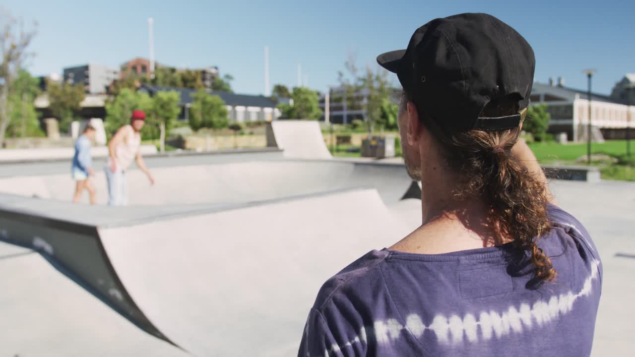 vista posterior del hombre caucásico en un día soleado, patinadores en el fondo