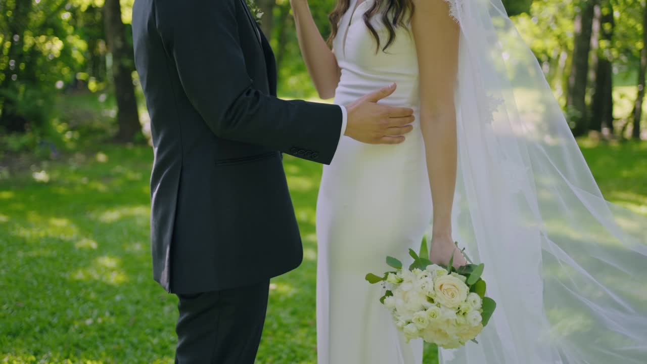 Bride holding a white rose bouquet as the groom embraces her in an outdoor setting